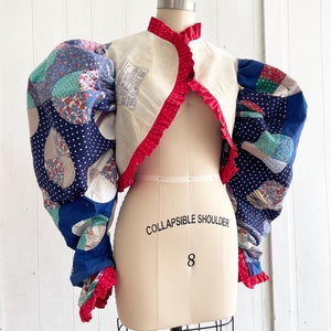 Puede incluir: Una chaqueta patchwork única con mangas abullonadas grandes. Las mangas presentan una mezcla de telas estampadas azules, blancas y rojas. La chaqueta tiene un ribete rojo con volantes y se exhibe en un maniquí con el texto "COLLAPSIBLE SHOULDER" y el número "8".