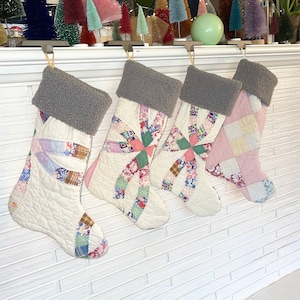 以下が含まれることがあります： 白いレンガの暖炉の棚に、グレーのシャーパの袖口が付いたキルティングされたクリスマスストッキングが4つぶら下がっています。ストッキングは、さまざまなカラフルなパッチワークパターンで飾られています。