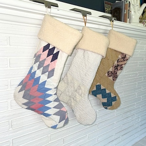 May include: Three patchwork fabric Christmas stockings hanging on a white brick fireplace mantel. The stockings are decorated with pink, blue, and gray geometric patterns.