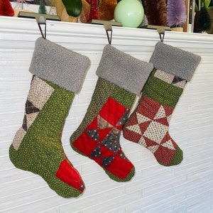 May include: Three green and red quilted Christmas stockings with gray sherpa cuffs hanging on a white brick fireplace mantel.