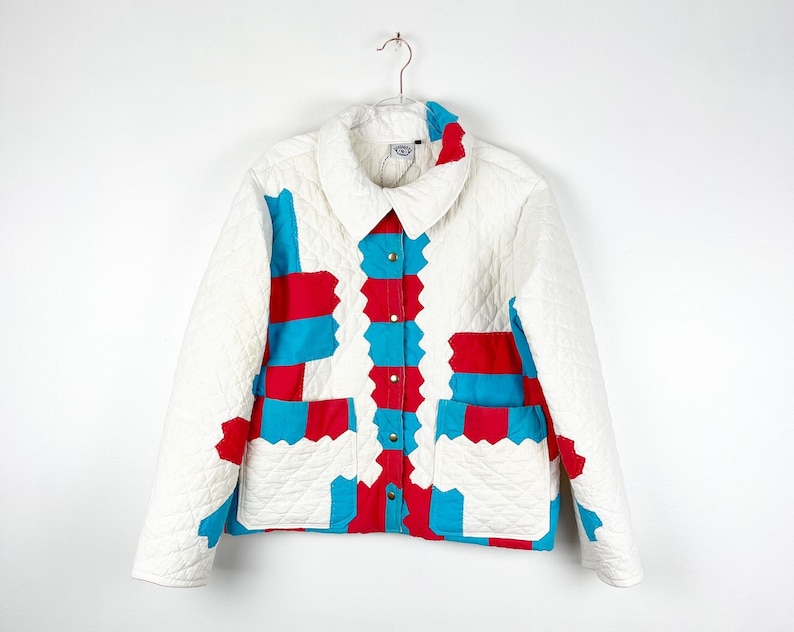 Puede incluir: Una chaqueta acolchada blanca con un dise&ntilde;o de patchwork azul y rojo. La chaqueta tiene cuello, cierre de botones y dos bolsillos.