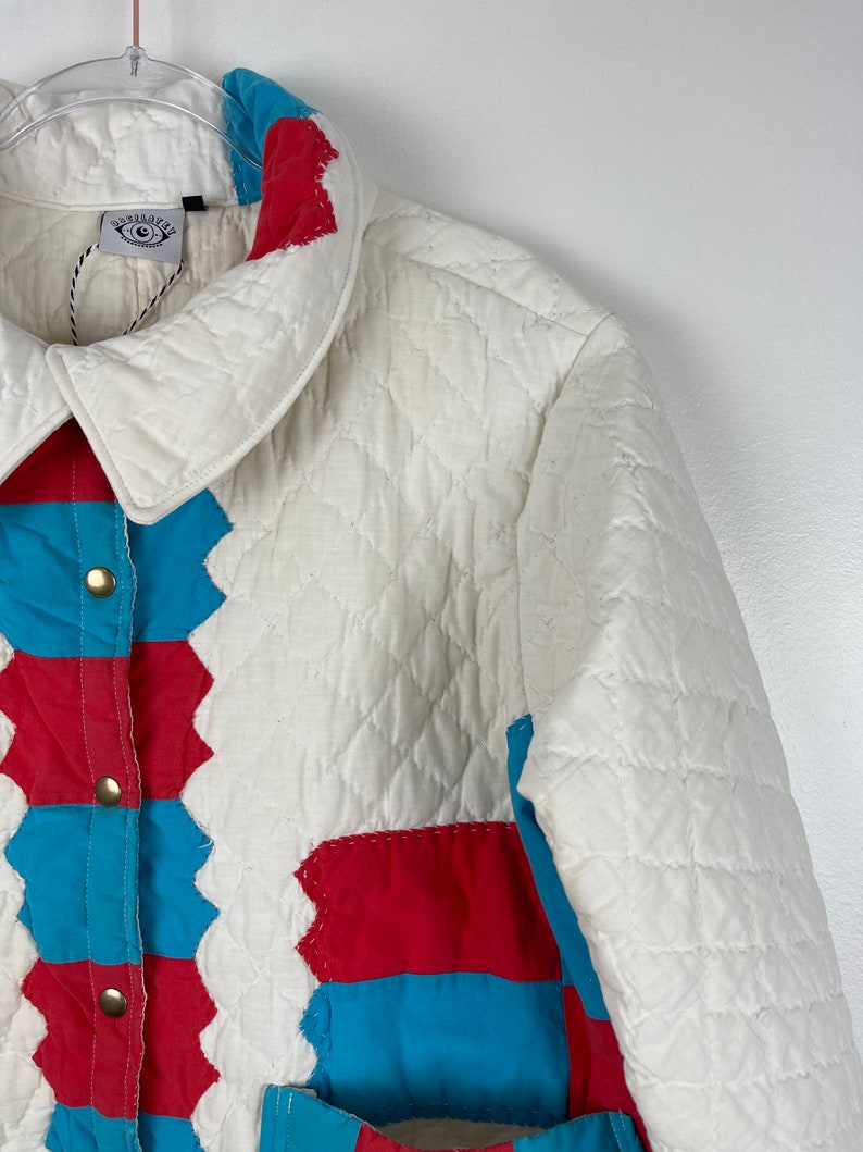 Puede incluir: Una chaqueta acolchada blanca con un dise&ntilde;o de patchwork con cuadrados rojos y azules. La chaqueta tiene un cierre de botones y un cuello.