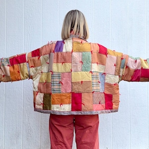 May include: A patchwork jacket in a variety of colours, including red, orange, yellow, and pink. The jacket is made of squares of fabric sewn together, with red bows at the seams. The sleeves have grey cuffs. The jacket is worn over red trousers.
