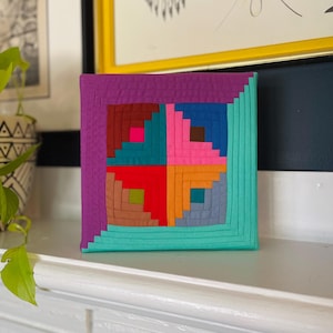 May include: A colorful square quilt block with a log cabin pattern. The block is made of fabric in shades of red, pink, blue, green, brown, and orange. The block is framed with a purple border.