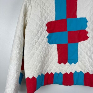 Puede incluir: Chaqueta acolchada blanca con un dise&ntilde;o de patchwork rojo y azul en la espalda. El patchwork tiene la forma de una cruz. La parte inferior de la chaqueta tiene un ribete a rayas rojo y azul.
