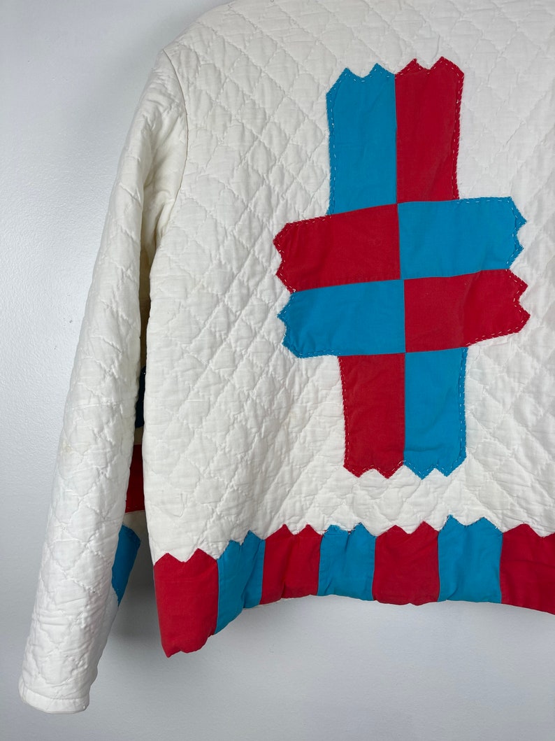 Puede incluir: Chaqueta acolchada blanca con un dise&ntilde;o de patchwork rojo y azul en la espalda. El patchwork tiene la forma de una cruz. La parte inferior de la chaqueta tiene un ribete a rayas rojo y azul.