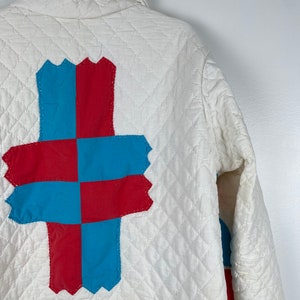 Puede incluir: Una chaqueta acolchada blanca con un dise&ntilde;o de patchwork rojo y azul en la espalda. El patchwork tiene la forma de una cruz.