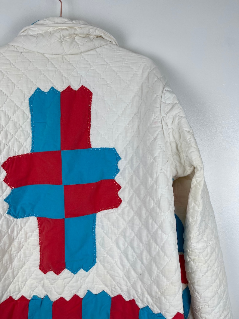 Puede incluir: Una chaqueta acolchada blanca con un dise&ntilde;o de patchwork rojo y azul en la espalda. El patchwork tiene la forma de una cruz.
