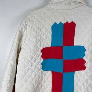 Puede incluir: Chaqueta acolchada blanca con un dise&ntilde;o de patchwork rojo y azul en la espalda. El patchwork tiene la forma de una cruz.