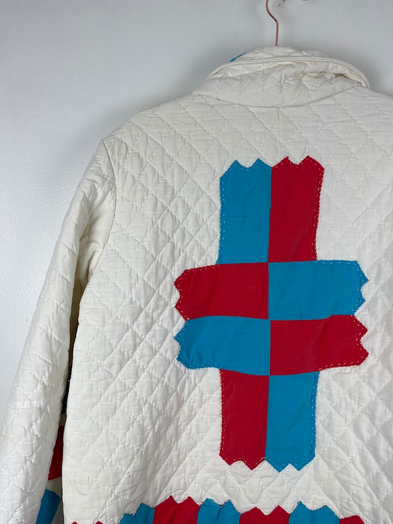 Puede incluir: Chaqueta acolchada blanca con un dise&ntilde;o de patchwork rojo y azul en la espalda. El patchwork tiene la forma de una cruz.