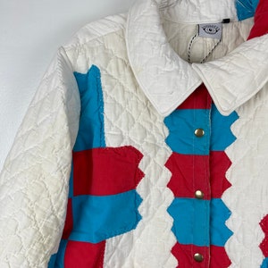 Puede incluir: Una chaqueta acolchada blanca con paneles de patchwork rojo y azul. La chaqueta tiene un cierre de botones y un cuello. La etiqueta de la chaqueta dice "Daydreamer".