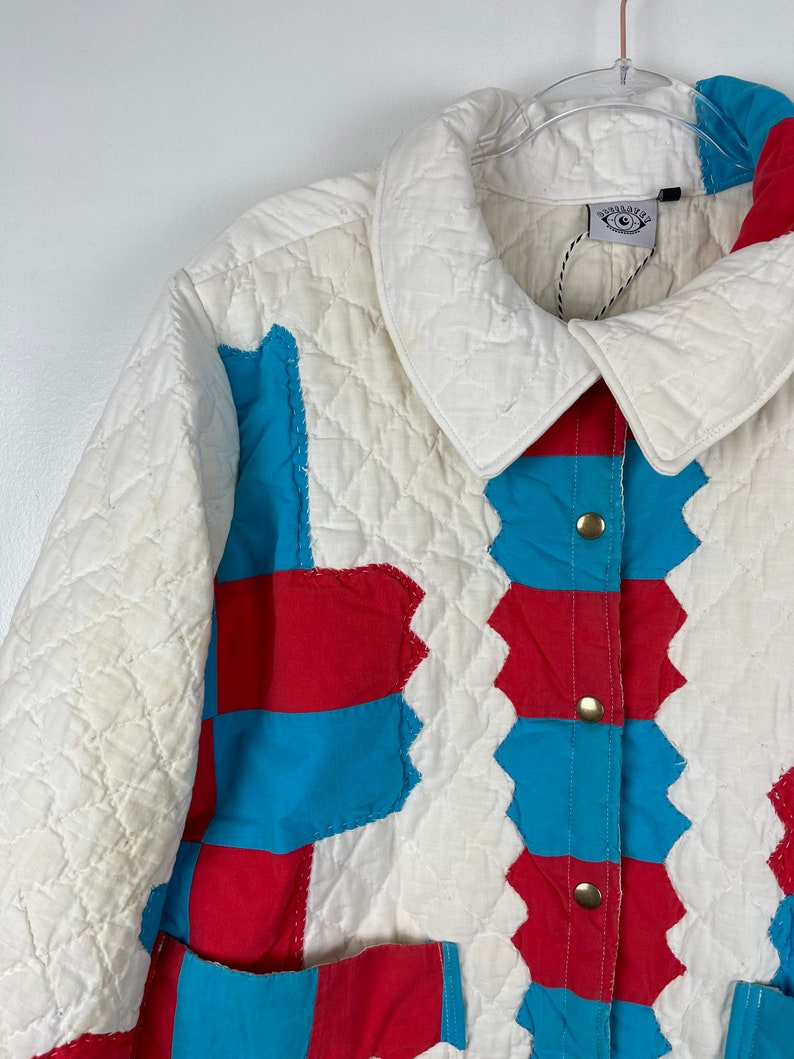 Puede incluir: Una chaqueta acolchada blanca con paneles de patchwork rojo y azul. La chaqueta tiene un cierre de botones y un cuello. La etiqueta de la chaqueta dice "Daydreamer".