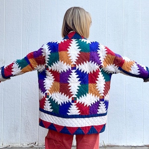 May include: A colorful, quilted jacket with a geometric pattern. The jacket features a patchwork design with squares of red, blue, green, purple, and orange. The jacket has long sleeves and a collar. The jacket is worn over red pants.