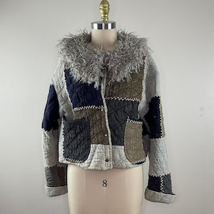 May include: A patchwork quilted jacket with a faux fur collar. The jacket is made of different fabrics in shades of blue, brown, and gray. The jacket has a button closure and two pockets.