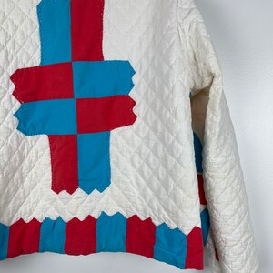 Puede incluir: Una chaqueta acolchada blanca con un dise&ntilde;o de patchwork azul y rojo en la espalda. El dise&ntilde;o presenta una forma de cruz con cuadrados alternos de tela azul y roja.