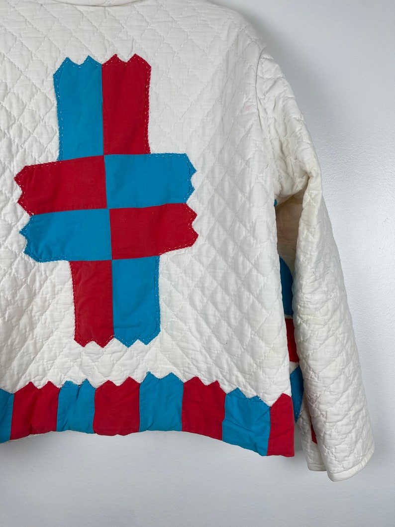 Puede incluir: Una chaqueta acolchada blanca con un dise&ntilde;o de patchwork azul y rojo en la espalda. El dise&ntilde;o presenta una forma de cruz con cuadrados alternos de tela azul y roja.