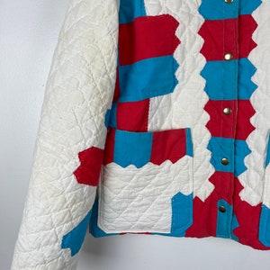 Puede incluir: Una chaqueta acolchada blanca con paneles de patchwork rojo y azul. La chaqueta tiene un cierre de botones en la parte delantera y un bolsillo de parche en el lado izquierdo.