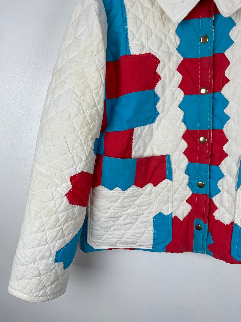 Puede incluir: Una chaqueta acolchada blanca con paneles de patchwork rojo y azul. La chaqueta tiene un cierre de botones en la parte delantera y un bolsillo de parche en el lado izquierdo.