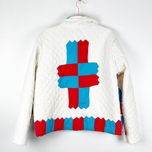Puede incluir: Chaqueta acolchada blanca con un dise&ntilde;o de patchwork rojo y azul en la espalda. El patchwork tiene la forma de una cruz.