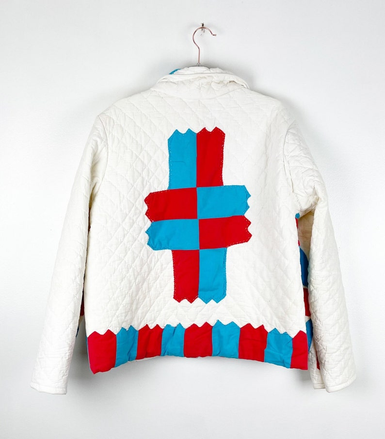 Puede incluir: Chaqueta acolchada blanca con un dise&ntilde;o de patchwork rojo y azul en la espalda. El patchwork tiene la forma de una cruz.