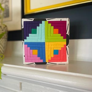 May include: A colorful fabric quilt block with a log cabin pattern in shades of pink, orange, yellow, green, blue, and purple. The block is set against a white background with black abstract lines.