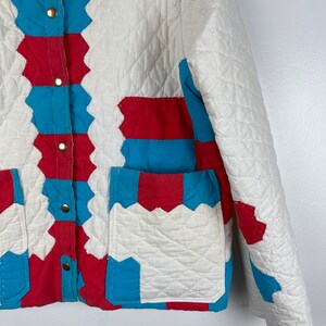 Puede incluir: Una chaqueta acolchada blanca con detalles de patchwork rojo y azul. La chaqueta tiene un cierre de botones y dos bolsillos de parche.