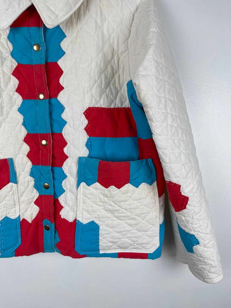 Puede incluir: Una chaqueta acolchada blanca con detalles de patchwork rojo y azul. La chaqueta tiene un cierre de botones y dos bolsillos de parche.