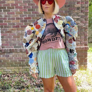 Puede incluir: Una mujer con un sombrero rosa, gafas de sol rojas en forma de corazón, una camiseta rosa con un estampado de armadillo negro y el texto "HOWDY!" impreso, una chaqueta corta floral multicolor y pantalones cortos de rayas verdes y blancas.