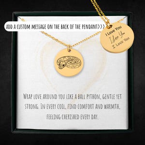 Op de afbeelding: Een goudkleurige hangerketting met een ronde bedel met een balpython-ontwerp. De ketting wordt gepresenteerd in een geschenkdoos met de tekst "I Love You" op een aparte bedel. De tekst "Voeg een aangepaste boodschap toe op de achterkant van de hanger" is ook zichtbaar.