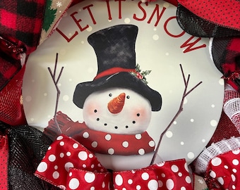 May include: A Christmas wreath with a snowman design at its centre. The snowman wears a black top hat and a red polka-dot scarf. The wreath features red and black plaid, polka-dot bows, and the text "LET IT SNOW".