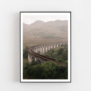 Può includere: Stampa incorniciata con un paesaggio panoramico di un viadotto ferroviario in pietra che si curva attraverso una valle. Il viadotto, con più archi, trasporta binari ferroviari su alberi e colline verdi sotto un cielo nebbioso. La cornice è nera.