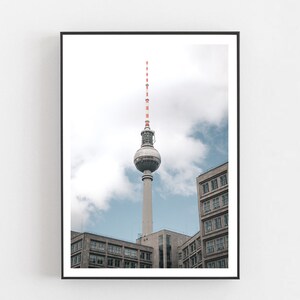 May include: A black and white photograph of the Berlin Television Tower, a tall, slender tower with a spherical observation deck, against a cloudy sky. The tower is surrounded by buildings.