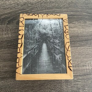 May include: A black and white photograph of a wooden bridge in a forest, framed in a natural wood frame with a burnt design around the edge.