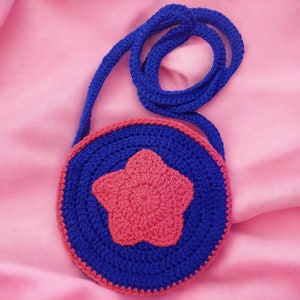 May include: A small, round crocheted purse with a blue and pink star design. The purse has a long strap for carrying over the shoulder.