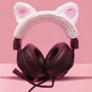 May include: Black over-ear headphones with a white crocheted cat ear headband. The cat ears have pink inner ear detailing. The headphones have a microphone and are set against a pink background.
