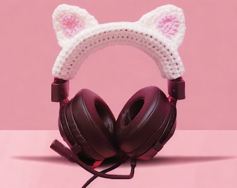 May include: Black over-ear headphones with a white crocheted cat ear headband. The cat ears have pink inner ear detailing. The headphones have a microphone and are set against a pink background.
