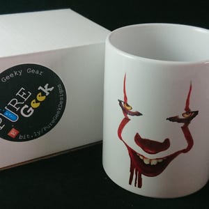 May include: White ceramic mug featuring a red and white clown face design. The mug is next to a white box with a black and white Pure Geek sticker. The mug has a handle and is likely for beverages.