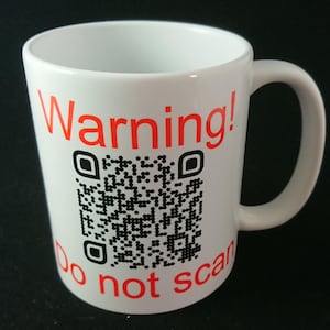 Op de afbeelding: Witte keramische mok met een rood waarschuwingsbord met de tekst "Waarschuwing! Niet scannen" en een zwart-witte QR-code.