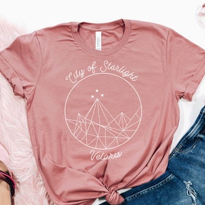 May include: A dusty rose t-shirt with a white graphic design that says "City of Starlight Velaris". The graphic features a geometric mountain range with stars inside a circle.