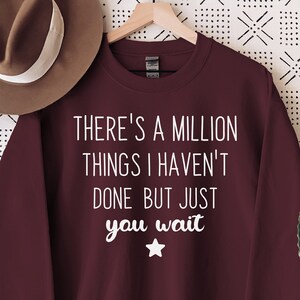 Peut inclure: Un sweat-shirt bordeaux avec le texte blanc "THERE'S A MILLION THINGS I HAVEN'T DONE BUT JUST YOU WAIT". Un chapeau marron est suspendu à gauche, et un cactus dans un pot blanc est posé sur une table en bois à droite. Le fond est un mur blanc à motifs.