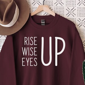 May include: A maroon sweatshirt with the words "RISE WISE EYES UP" in white lettering. A brown hat hangs to the left, and a cactus in a white pot sits on a wooden table to the right. The background is a white wall with a black geometric pattern.
