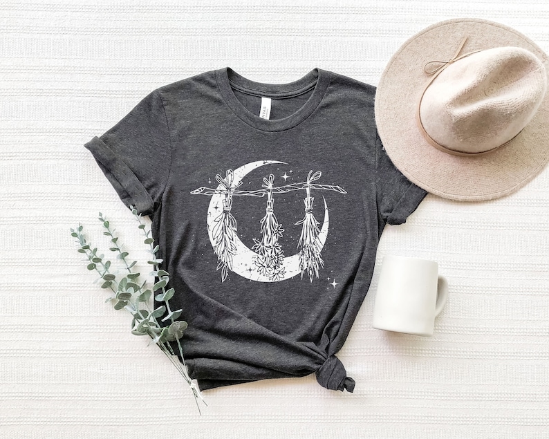 Pu&ograve; includere: T-shirt grigio scuro con grafica bianca a forma di luna crescente e mazzi di erbe appesi. La maglietta &egrave; abbinata a un cappello beige e una tazza bianca. La maglietta &egrave; annodata in vita.