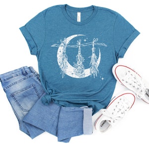Pu&ograve; includere: T-shirt color teal con un design a mezzaluna bianca e erbe appese. La maglietta &egrave; abbinata a jeans blu e scarpe da ginnastica di tela bianche. Il design presenta elementi botanici e stelle.