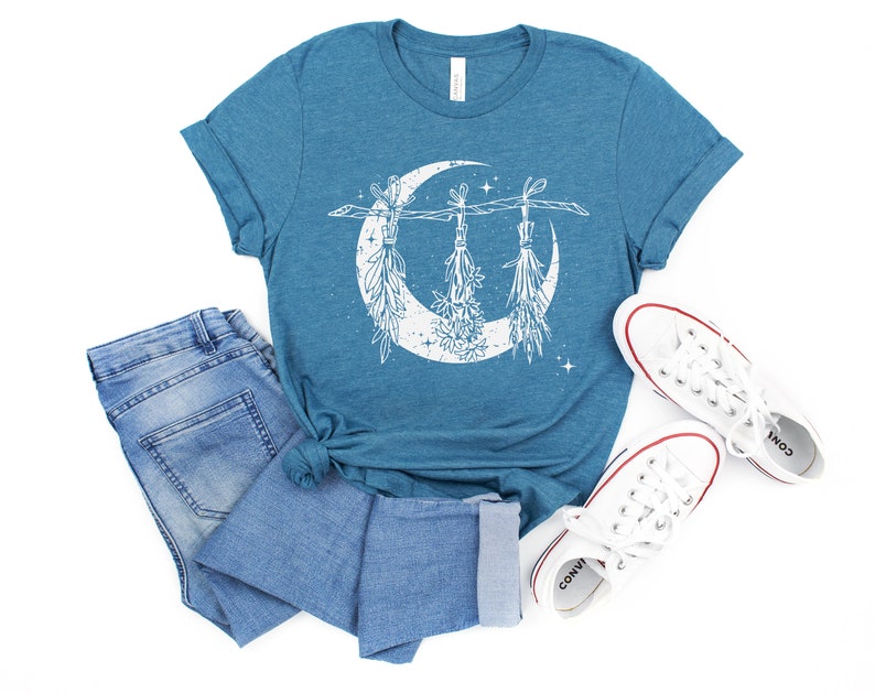 Pu&ograve; includere: T-shirt color teal con un design a mezzaluna bianca e erbe appese. La maglietta &egrave; abbinata a jeans blu e scarpe da ginnastica di tela bianche. Il design presenta elementi botanici e stelle.
