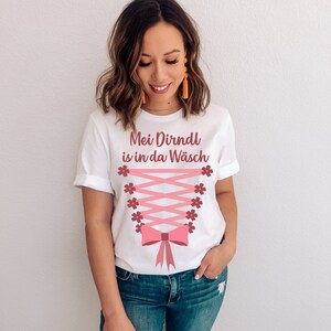 Peut inclure: T-shirt blanc avec le texte "Mei Dirndl is in da W&auml;sch" en lettres roses. Le t-shirt pr&eacute;sente un design de corset rose avec un n&oelig;ud et des accents floraux.