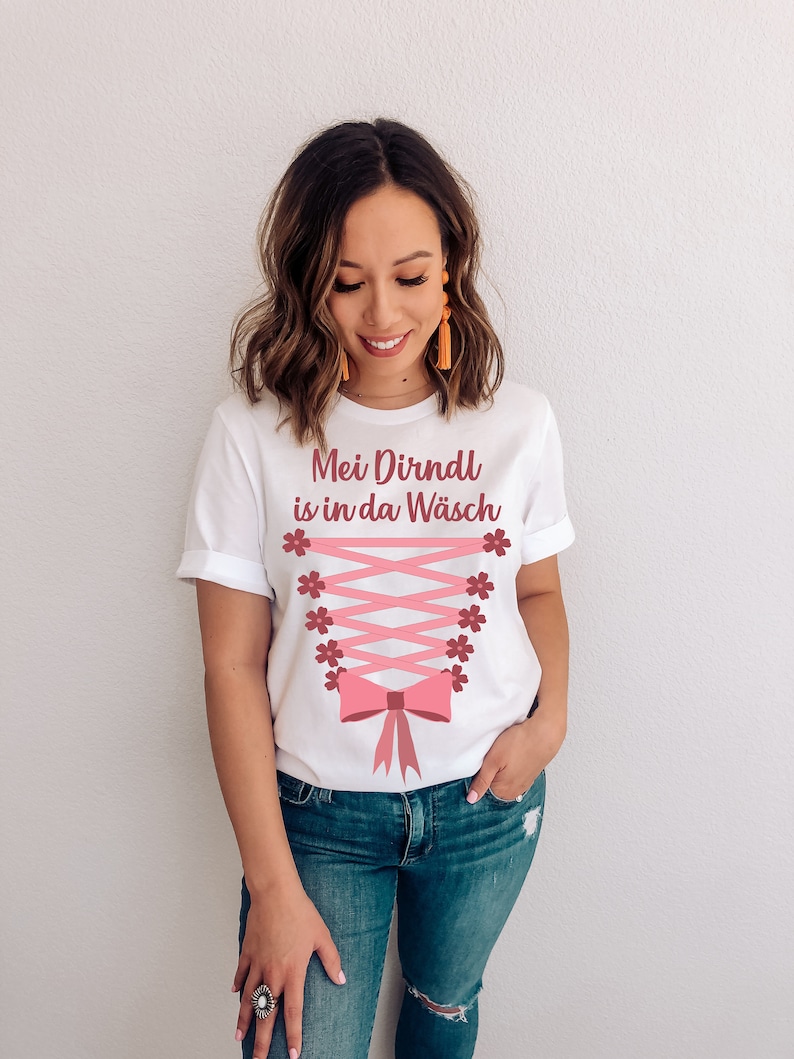 Peut inclure: T-shirt blanc avec le texte "Mei Dirndl is in da W&auml;sch" en lettres roses. Le t-shirt pr&eacute;sente un design de corset rose avec un n&oelig;ud et des accents floraux.