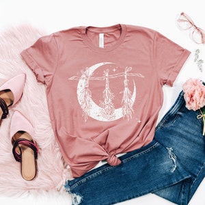 Pu&ograve; includere: Una maglietta rosa antico con una grafica bianca di una luna crescente con mazzi di erbe appesi. La maglietta &egrave; abbinata a jeans blu, muli rosa e un tappeto in finta pelliccia rosa. Un bouquet di fiori rosa e occhiali completano l'estetica.