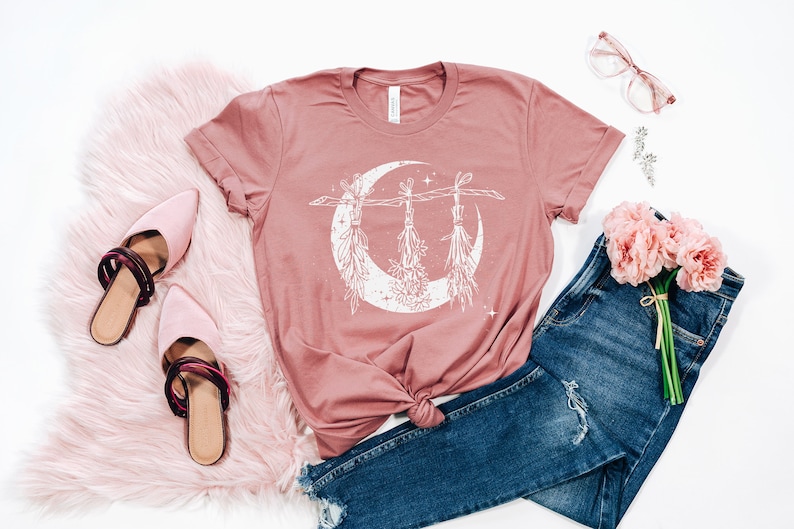 Pu&ograve; includere: Una maglietta rosa antico con una grafica bianca di una luna crescente con mazzi di erbe appesi. La maglietta &egrave; abbinata a jeans blu, muli rosa e un tappeto in finta pelliccia rosa. Un bouquet di fiori rosa e occhiali completano l'estetica.
