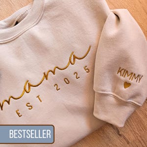 Könnte beinhalten: Ein hellbeiger Pullover mit dem Schriftzug "mama est 2025" in Gold auf der Vorderseite und dem Schriftzug "KIMMY" mit einem kleinen Herz in Gold auf dem Ärmel gestickt.