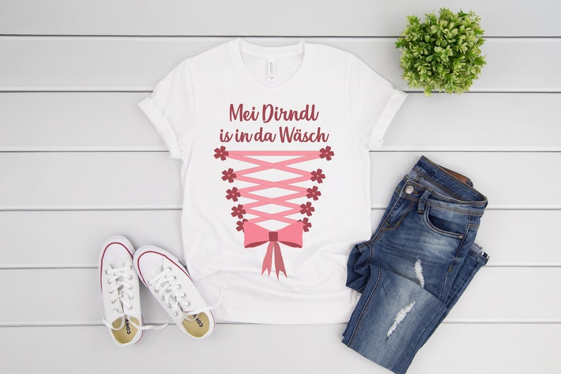 Peut inclure: T-shirt blanc avec le texte "Mei Dirndl is in da W&auml;sch" en lettres rouges. Le t-shirt pr&eacute;sente un design de corset rose avec un n&oelig;ud en bas.